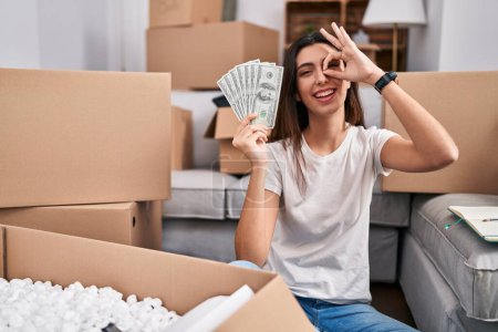 Photo for Young beautiful woman sitting on the floor at new home holding money smiling happy doing ok sign with hand on eye looking through fingers - Royalty Free Image