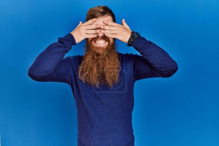 Foto de Redhead man with long beard wearing casual blue sweater over blue background covering eyes with hands smiling cheerful and funny. blind concept. - Imagen libre de derechos