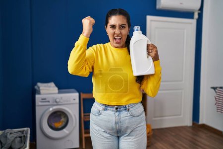 Téléchargez les photos : Young hispanic woman holding detergent bottle annoyed and frustrated shouting with anger, yelling crazy with anger and hand raised - en image libre de droit