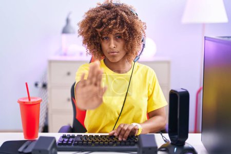 Foto de Young hispanic woman with curly hair playing video games wearing headphones doing stop sing with palm of the hand. warning expression with negative and serious gesture on the face. - Imagen libre de derechos