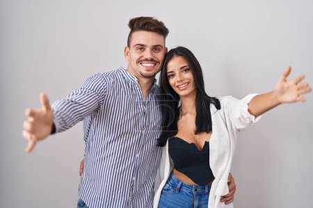 Photo for Young hispanic couple standing over white background looking at the camera smiling with open arms for hug. cheerful expression embracing happiness. - Royalty Free Image