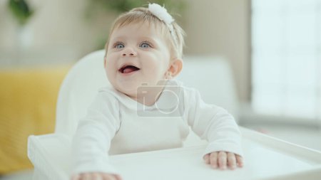 Foto de Adorable bebé rubio sonriendo confiado sentado en la trona en casa - Imagen libre de derechos