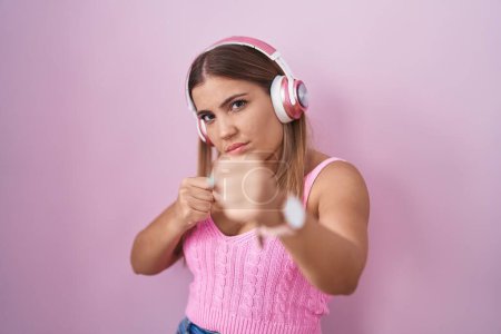 Photo for Young blonde woman listening to music using headphones ready to fight with fist defense gesture, angry and upset face, afraid of problem - Royalty Free Image