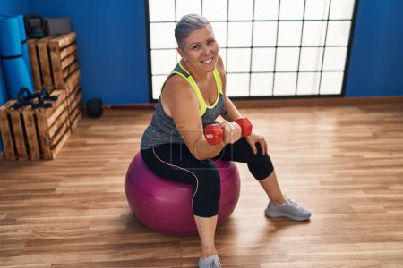 Photo for Middle age woman training with dumbbell sitting on fit ball at sport center - Royalty Free Image