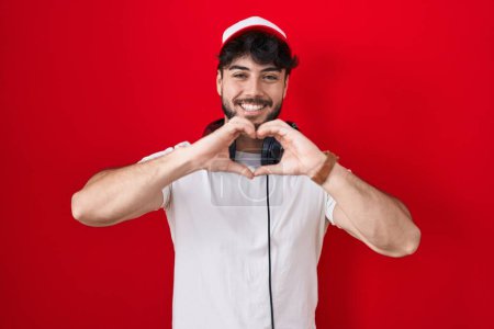 Téléchargez les photos : Hispanic man with beard wearing gamer hat and headphones smiling in love doing heart symbol shape with hands. romantic concept. - en image libre de droit