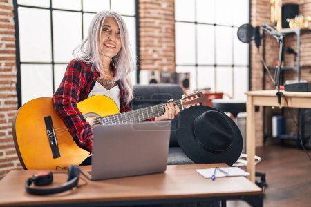 Téléchargez les photos : Femme musicienne aux cheveux gris du Moyen Âge jouant de la guitare classique avec un ordinateur portable dans un studio de musique - en image libre de droit