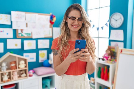 Téléchargez les photos : Jeune enseignante blonde utilisant un smartphone debout à la maternelle - en image libre de droit