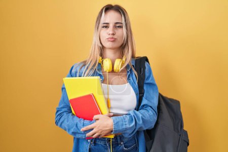 Foto de Mujer rubia joven con mochila de estudiante y sosteniendo libros hinchando mejillas con cara divertida. boca hinchada de aire, expresión loca. - Imagen libre de derechos