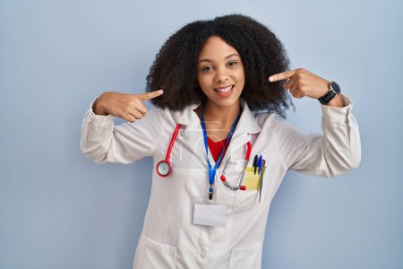 Téléchargez les photos : Jeune femme afro-américaine portant l'uniforme de médecin et stéthoscope souriant joyeuse montrant et pointant du doigt les dents et la bouche. concept de santé dentaire. - en image libre de droit