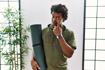 Photo for African man with curly hair holding yoga mat at studio pointing to the eye watching you gesture, suspicious expression - Royalty Free Image