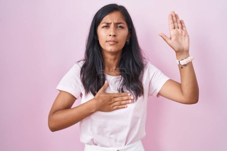 Photo for Young hispanic woman standing over pink background swearing with hand on chest and open palm, making a loyalty promise oath - Royalty Free Image