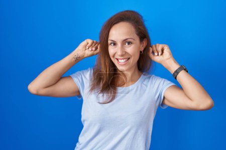Téléchargez les photos : Femme brune debout sur fond bleu souriant tirant les oreilles avec les doigts, geste drôle. problème d'audition - en image libre de droit