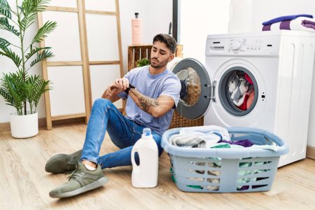 Téléchargez les photos : Young hispanic man putting dirty laundry into washing machine checking the time on wrist watch, relaxed and confident - en image libre de droit