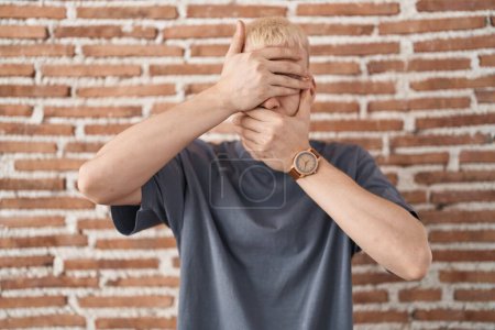 Foto de Young caucasian man standing over bricks wall covering eyes and mouth with hands, surprised and shocked. hiding emotion - Imagen libre de derechos