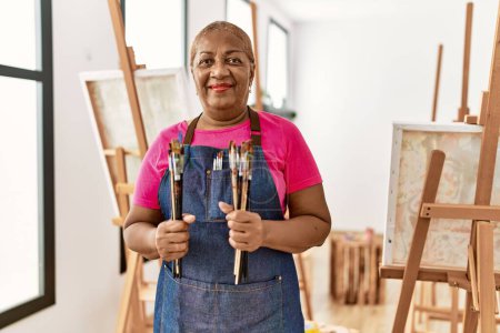 Foto de Mujer afroamericana mayor sonriendo confiada sosteniendo pinceles en el estudio de arte - Imagen libre de derechos