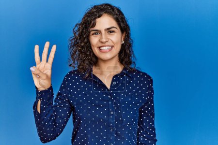 Photo for Young brunette woman with curly hair wearing casual clothes over blue background showing and pointing up with fingers number three while smiling confident and happy. - Royalty Free Image