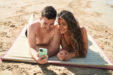 Foto de Jóvenes turistas hispanos en pareja que usan traje de baño con smartphone en la playa - Imagen libre de derechos