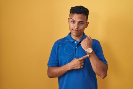 Téléchargez les photos : Jeune homme hispanique debout sur fond jaune pressé pointant vers regarder le temps, l'impatience, regardant la caméra avec une expression détendue - en image libre de droit