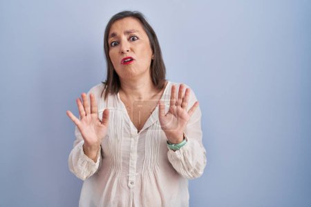 Foto de Middle age hispanic woman standing over blue background moving away hands palms showing refusal and denial with afraid and disgusting expression. stop and forbidden. - Imagen libre de derechos