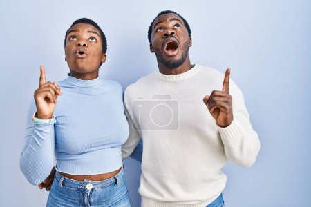 Photo for Young african american couple standing over blue background amazed and surprised looking up and pointing with fingers and raised arms. - Royalty Free Image