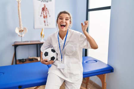 Téléchargez les photos : Jeune femme travaillant à la clinique de thérapie de football célébrant la victoire avec sourire heureux et expression gagnante avec les mains levées - en image libre de droit