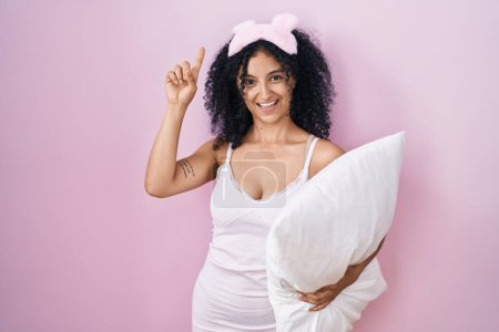 Photo for Hispanic woman with curly hair wearing sleep mask and pajama holding pillow pointing finger up with successful idea. exited and happy. number one. - Royalty Free Image