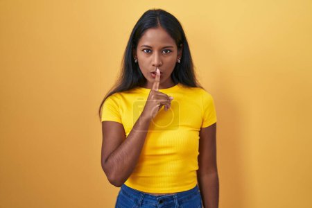 Photo for Young indian woman standing over yellow background asking to be quiet with finger on lips. silence and secret concept. - Royalty Free Image