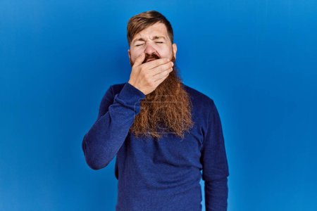 Photo for Redhead man with long beard wearing casual blue sweater over blue background bored yawning tired covering mouth with hand. restless and sleepiness. - Royalty Free Image