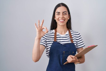 Foto de Young brunette woman wearing professional waitress apron and clipboard doing ok sign with fingers, smiling friendly gesturing excellent symbol - Imagen libre de derechos