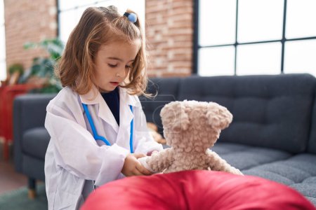 Photo for Adorable hispanic girl wearing doctor uniform putting band aid on teddy bear arm at home - Royalty Free Image