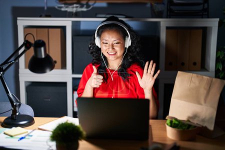 Foto de Joven mujer asiática trabajando en la oficina con el ordenador portátil en la noche mostrando y apuntando hacia arriba con los dedos número seis mientras sonríe confiado y feliz. - Imagen libre de derechos