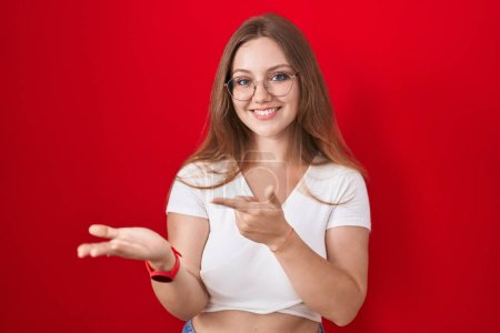 Photo for Young caucasian woman standing over red background amazed and smiling to the camera while presenting with hand and pointing with finger. - Royalty Free Image