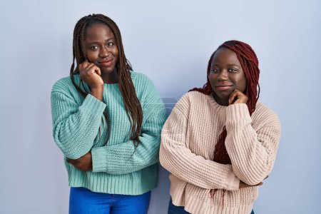 Téléchargez les photos : Deux femmes africaines debout sur fond bleu avec la main sur le menton penser à la question, expression coûteuse. souriant avec un visage réfléchi. concept de doute. - en image libre de droit