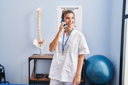 Foto de Mujer rubia joven con uniforme de fisioterapeuta hablando en el teléfono inteligente en la clínica - Imagen libre de derechos