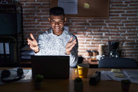 Téléchargez les photos : Young hispanic man working at the office at night looking at the camera smiling with open arms for hug. cheerful expression embracing happiness. - en image libre de droit