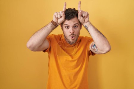 Téléchargez les photos : Young hispanic man standing over yellow background doing funny gesture with finger over head as bull horns - en image libre de droit