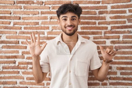 Foto de Hombre árabe con barba de pie sobre ladrillos fondo de la pared mostrando y apuntando hacia arriba con los dedos número seis mientras sonríe confiado y feliz. - Imagen libre de derechos
