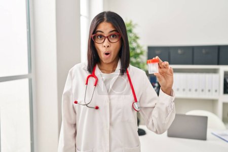 Foto de Young hispanic doctor woman holding sample cup scared and amazed with open mouth for surprise, disbelief face - Imagen libre de derechos