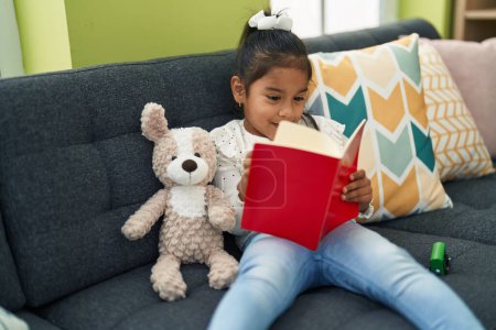 Téléchargez les photos : Adorable hispanique fille lecture livre assis sur le canapé à la maison - en image libre de droit