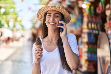 Foto de Young hispanic woman tourist talking on the smartphone eating ice cream at street market - Imagen libre de derechos