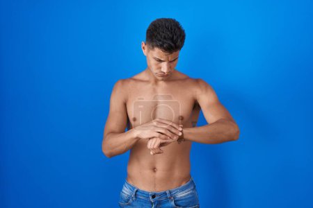 Foto de Young hispanic man standing shirtless over blue background checking the time on wrist watch, relaxed and confident - Imagen libre de derechos