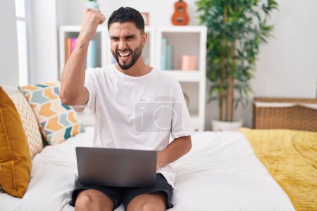 Foto de Hispanic young man using computer laptop sitting on the bed annoyed and frustrated shouting with anger, yelling crazy with anger and hand raised - Imagen libre de derechos
