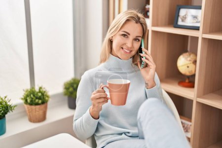 Photo for Young blonde woman talking on the smartphone drinking coffee at home - Royalty Free Image