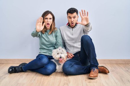 Photo for Young hispanic couple sitting on the floor with dog doing stop gesture with hands palms, angry and frustration expression - Royalty Free Image