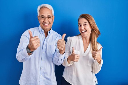 Foto de Middle age hispanic couple standing over blue background success sign doing positive gesture with hand, thumbs up smiling and happy. cheerful expression and winner gesture. - Imagen libre de derechos