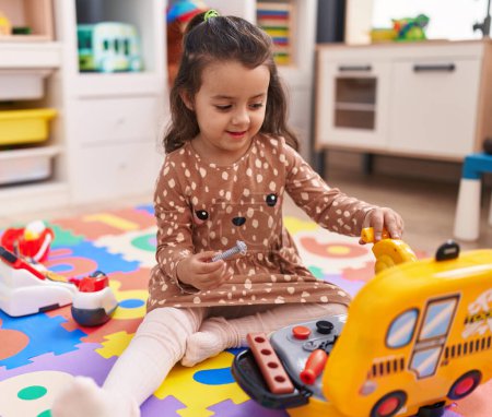 Foto de Adorable chica hispana sentada en el suelo jugando con un juguete mecánico en el jardín de infantes - Imagen libre de derechos
