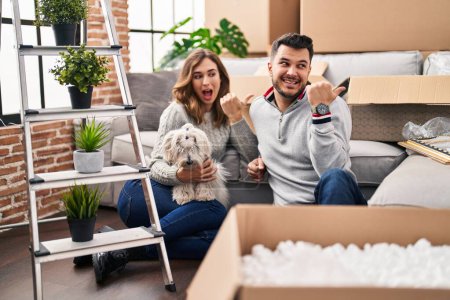 Photo for Young hispanic couple sitting on the floor at new home with log pointing thumb up to the side smiling happy with open mouth - Royalty Free Image