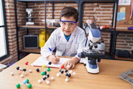 Foto de Adorable hispanic boy student using microscope writing on notebook at laboratory classroom - Imagen libre de derechos