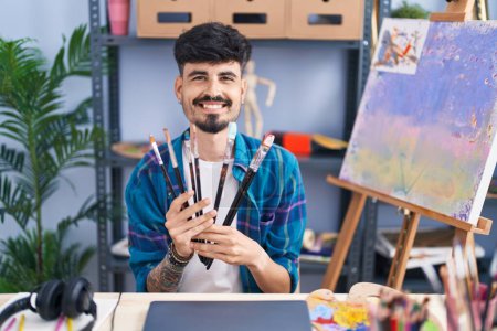 Young hispanic man artist smiling confident holding paintbrushes at art studio