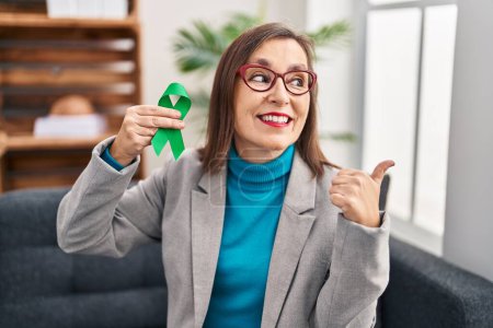 Photo for Middle age hispanic woman holding support green ribbon pointing thumb up to the side smiling happy with open mouth - Royalty Free Image